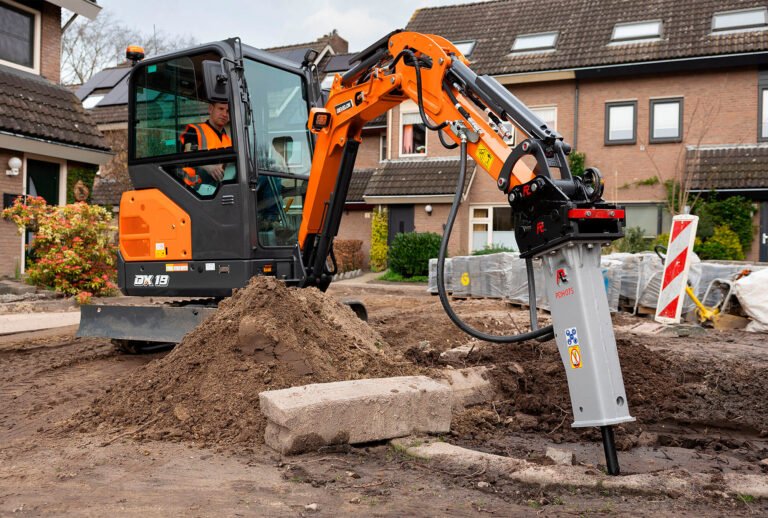 Construction worker operating a mini excavator with a hydraulic breaker attachment to break concrete and prepare the ground on a residential site.