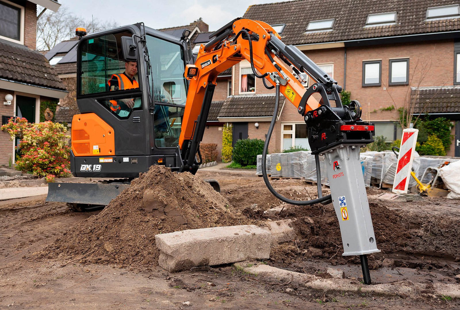 Construction worker operating a mini excavator with a hydraulic breaker attachment to break concrete and prepare the ground on a residential site.